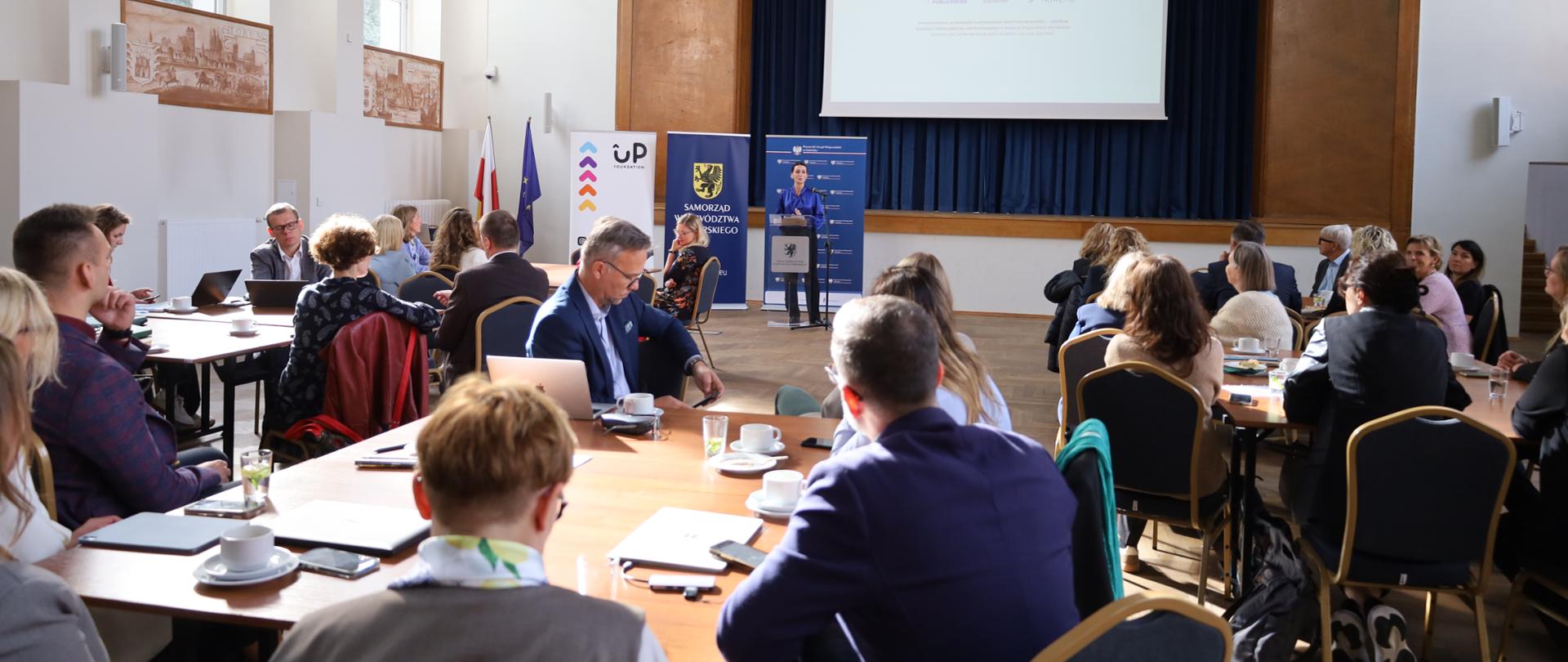 Na zdjęciu sala i kilka stołów, przy których siedzą ludzie. Przy mównicy stoi kobieta (wojewoda pomorska).