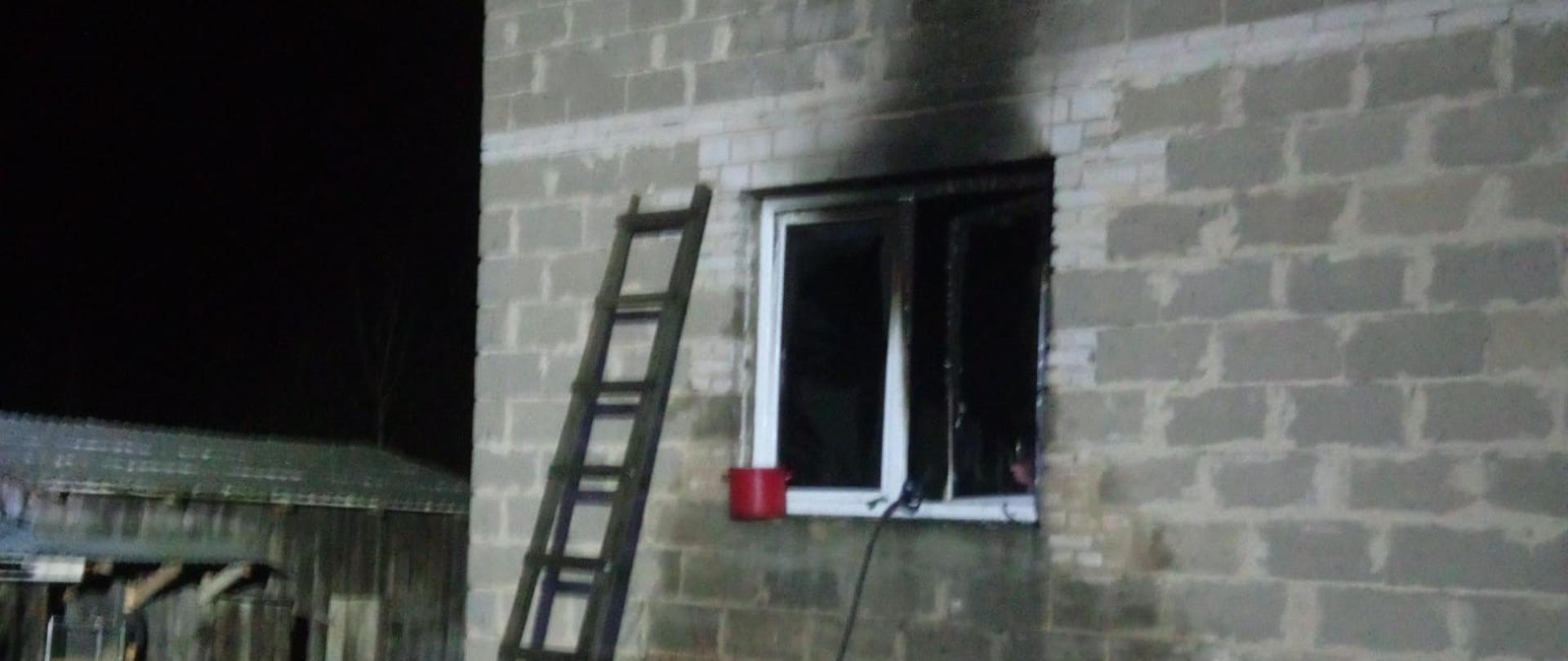 Zdjęcie przedstawia budynek murowany mieszkalny gdzie doszło do pożaru. Okno na parterze jest mocno okopcone i widać przystawioną do ściany drabinę.