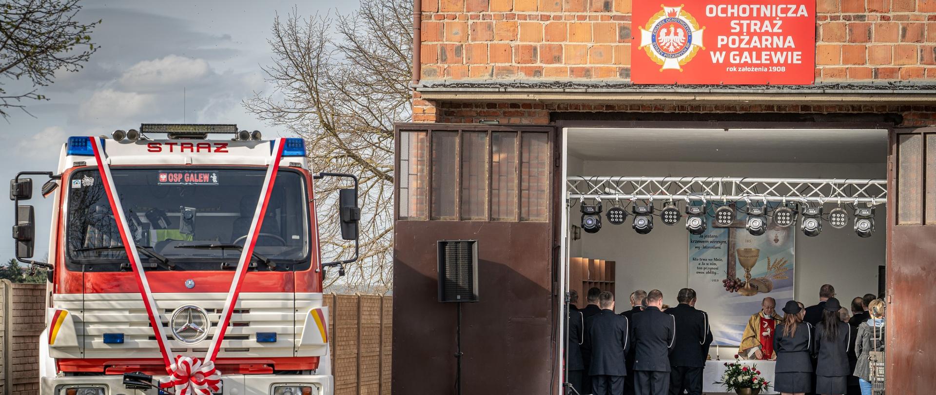 Po lewej stronie widoczny czerwony samochód strażacki ozdobiony wstęgą, po prawie ceglana remiza z otwartymi drzwiami garazowymi, w remizie widoczny ksiądz odprawiający mszę i stojace osoby w mundurach wyjściowych strażacy pożarnych