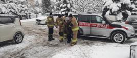 Kolorowe zdjęcie przedstawia na środku, bok srebrnego samochodu pożarniczego Państwowej Straży Pożarnej z napisem Straż na czerwonym pasem. Obok pojazdu czterej umundurowani strażacy Państwowej Straży Pożarnej i Ochotniczej Straży Pożarnej pomagają starszej osobie wyjść z pojazdu. Na całym planie fotografii widoczne mocno zaśnieżone pojazdy osobowe na parkingu i duża ilość śniegu. W tle zaśnieżone drzewa.
