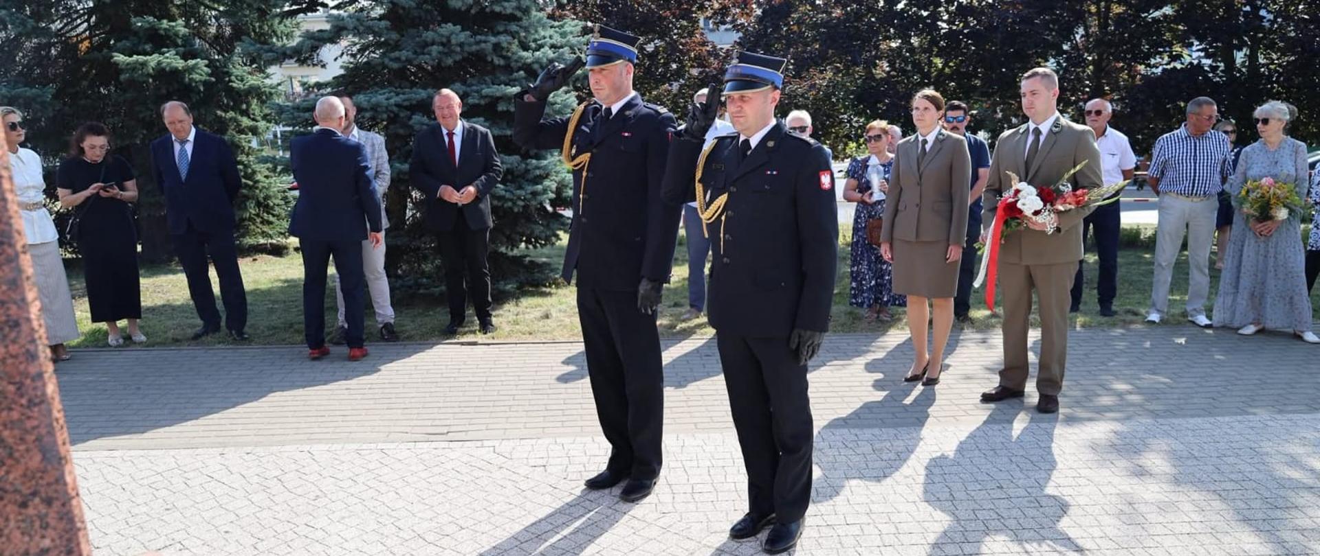 Uroczystość patriotyczna – delegacje i mieszkańcy stoją przed pomnikiem, dwóch strażaków w galowych mundurach oddaje salut, a pozostali uczestnicy trzymają kwiaty i znicze.