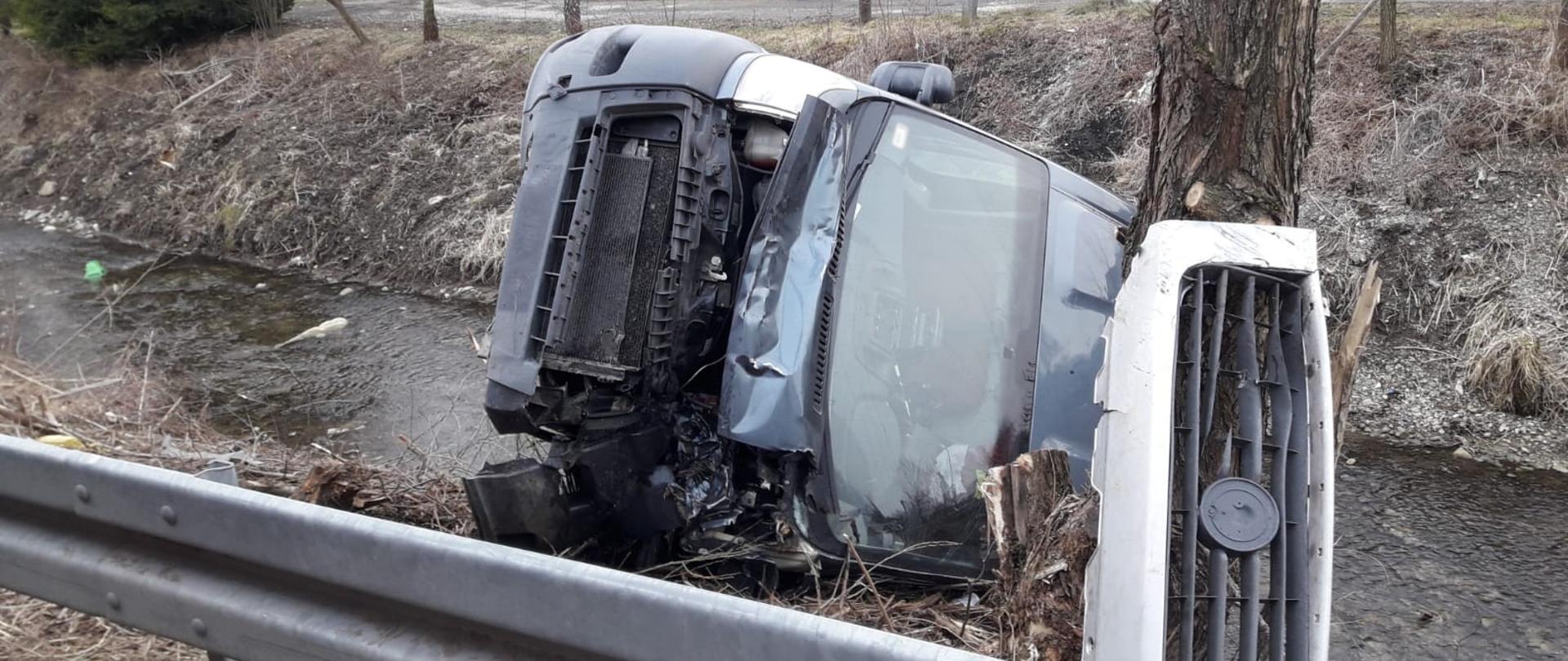 Samochód Fiat Ducato leży na lewym boku na brzegu potoku obok drzewa. W oddali radiowóz Policji obok którego stoją ludzie a także budynek.