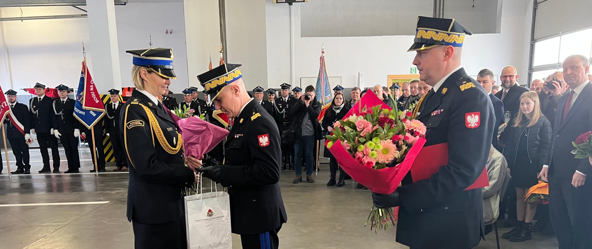 strażak wręcza kwiaty oraz prezent kobiecie strażak, za nim stoi drugi strażak z kwaitali, w tle gruupa strażaków