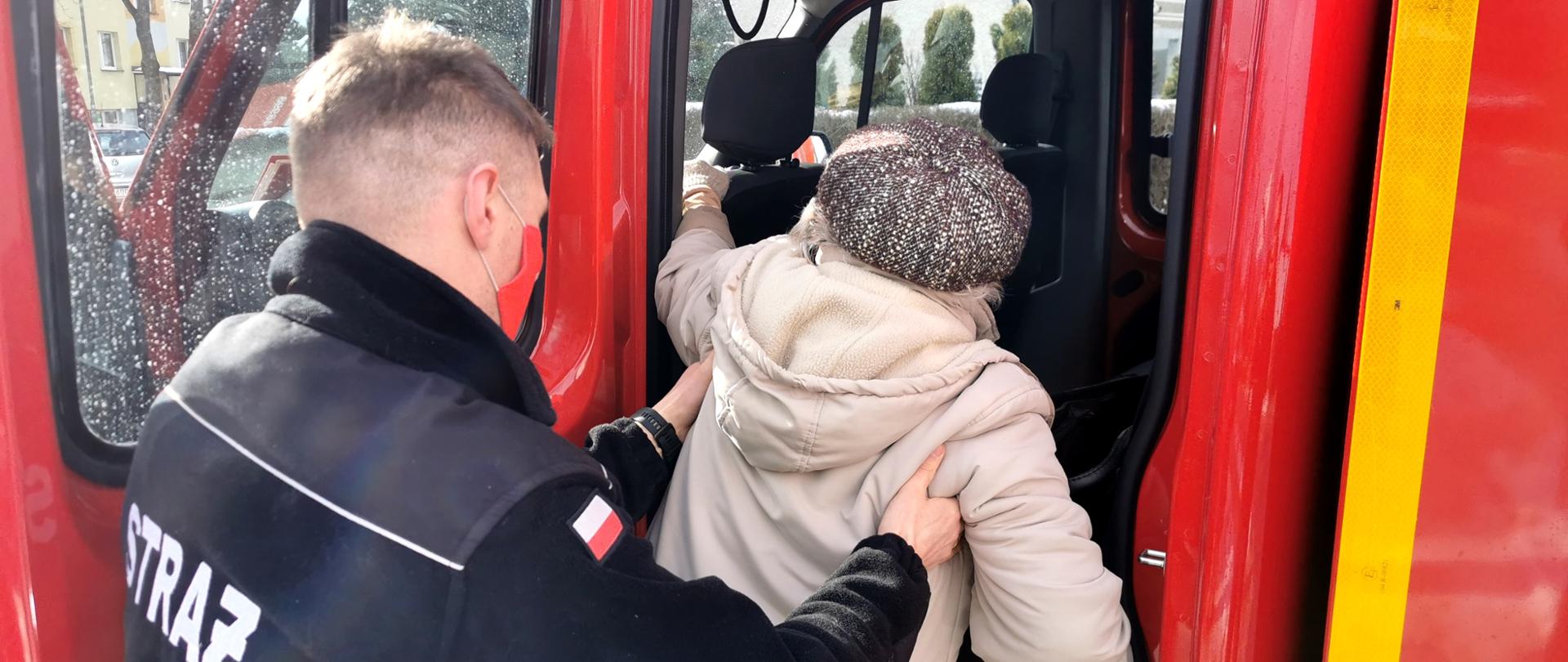 Strażak Ochotniczej Straży Pożarnej pomaga starszej pani wejść do samochodu pożarniczego