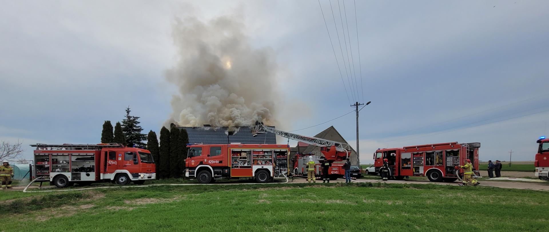 Na zdjęciu widać 5 samochodów pożarniczych. W tle nad budynkiem mieszkalnym widać kłęby dymu z palącego się poddasza domu jednorodzinnego