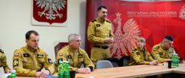 Na zdjęciu przedstawiona jest grupa strażaków Państwowej Straży Pożarnej (PSP) uczestniczących w szkoleniu w sali konferencyjnej. Mężczyzna w beżowym mundurze, stojący przy stole, przemawia do zgromadzonych. Jego mundur ma naszywkę z polską flagą oraz oznaczenia stopnia służbowego.
Pozostali strażacy siedzą przy dużym drewnianym stole, słuchając wypowiedzi. Na stole znajdują się butelki z wodą, notatniki oraz drobne przekąski. W tle widać ścianę ozdobioną czerwonym banerem z grafiką białego orła oraz napisem „PAŃSTWOWA STRAŻ POŻARNA KOMENDA MIEJSKA PAŃSTWOWEJ STRAŻY POŻARNEJ W TARNOWIE”. Na ścianie powieszony jest również herb Polski – biały orzeł w koronie na czerwonym tle – oraz krzyż.