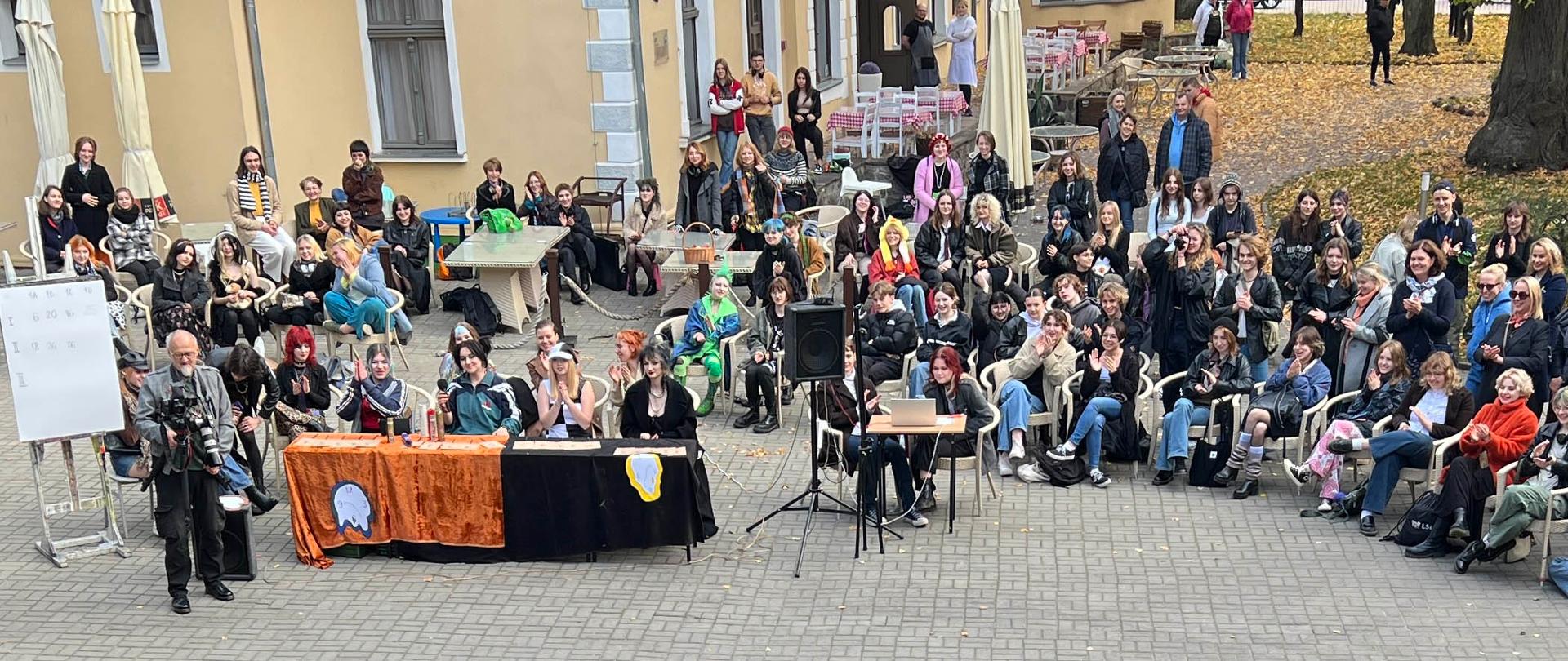 Zabawa otrzęsin na dziedzińcu szkoły. Grupa uczniów i nauczycieli przygląda się zabawie.