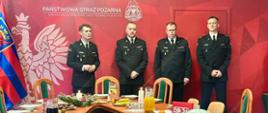 Cztery osoby w mundurach strażackich stoją za stołem konferencyjnym ozdobionym świątecznymi dekoracjami i nakryciami, w pomieszczeniu z flagą i banerem Komendy Miejskiej Państwowej Straży Pożarnej w Szczecinie.
