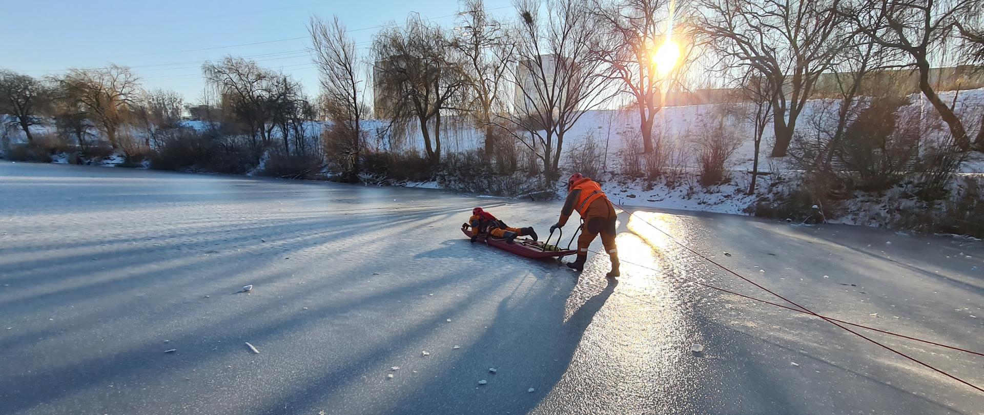 Zdjęcie przedstawia strażaków ćwiczących na lodzie. Jeden ze strażaków znajduje się na desce lodowej, drugi pcha deskę w celu dotarcia do poszkodowanego. 