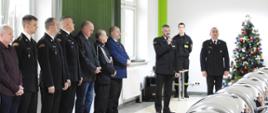 Zdjęcie przedstawia strażaków i pracowników Komendy podczas spotkania opłatkowego w Komendzie Powiatowej Państwowej Straży Pożarnej w Tomaszowie Mazowieckim. Głos zabiera zastępca łódzkiego komendanta wojewódzkiego PSP w Łodzi - starszy brygadier Paweł Malinowski. Po jego prawej stronie stoi komendant powiatowy PSP w Tomaszowie Mazowieckim. Za nim jest przystrojona choinka.