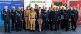 Strażacy oraz przedstawiciele funduszu ochrony środowiska na wspólnym zdjęciu w sali, w której wręczano umowy na dofinansowanie, w tle telewizor z wyświetlaną prezentacją. 