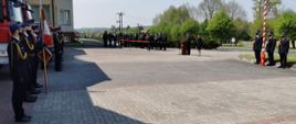 Zdjęcie przedstawia plac przed budynkiem komendy, wyłożony kostką. Po lewej stronie stoją w dwuszeregu strażacy w galowym umundurowaniu, a za nimi stoją samochody strażackie. Na wprost widać stojących zaproszonych gości, a po prawo znajduje się mównica, przy której stoją dwie osoby, mężczyzna i kobieta w galowym umundurowaniu, a następnie masz z flagą i stojący przy niej trzej strażacy w galowym umundurowaniu.