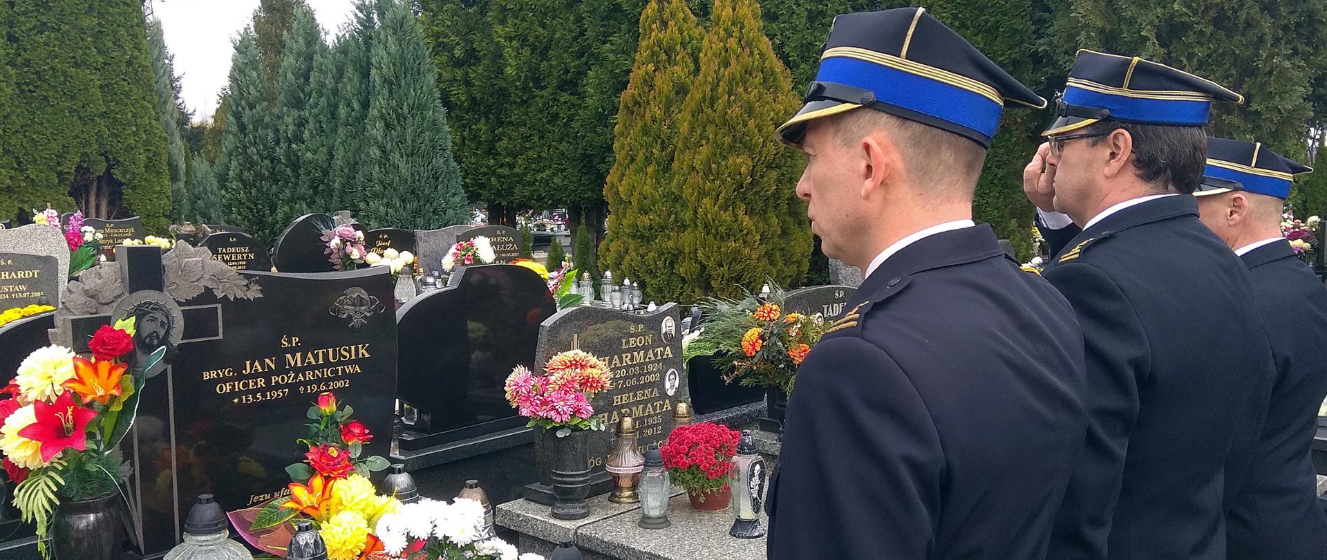 Widok z tyłu. Na cmentarzu przed grobem stoją trzej strażacy w mundurach wyjściowych i salutują. 