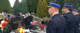 Widok z tyłu. Na cmentarzu przed grobem stoją trzej strażacy w mundurach wyjściowych i salutują. 
