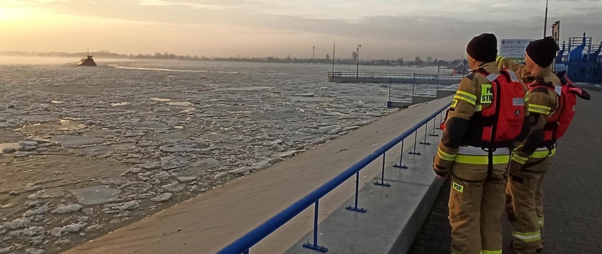 Strażacy patrolują wały. Dwóch strażaków w ubraniach specjalnych i czerwonych kamizelkach asekuracyjnych stoi na betonowym wale patrząc na zamarzniętą rzekę Wisłę. W tle na rzece lodołamacz.