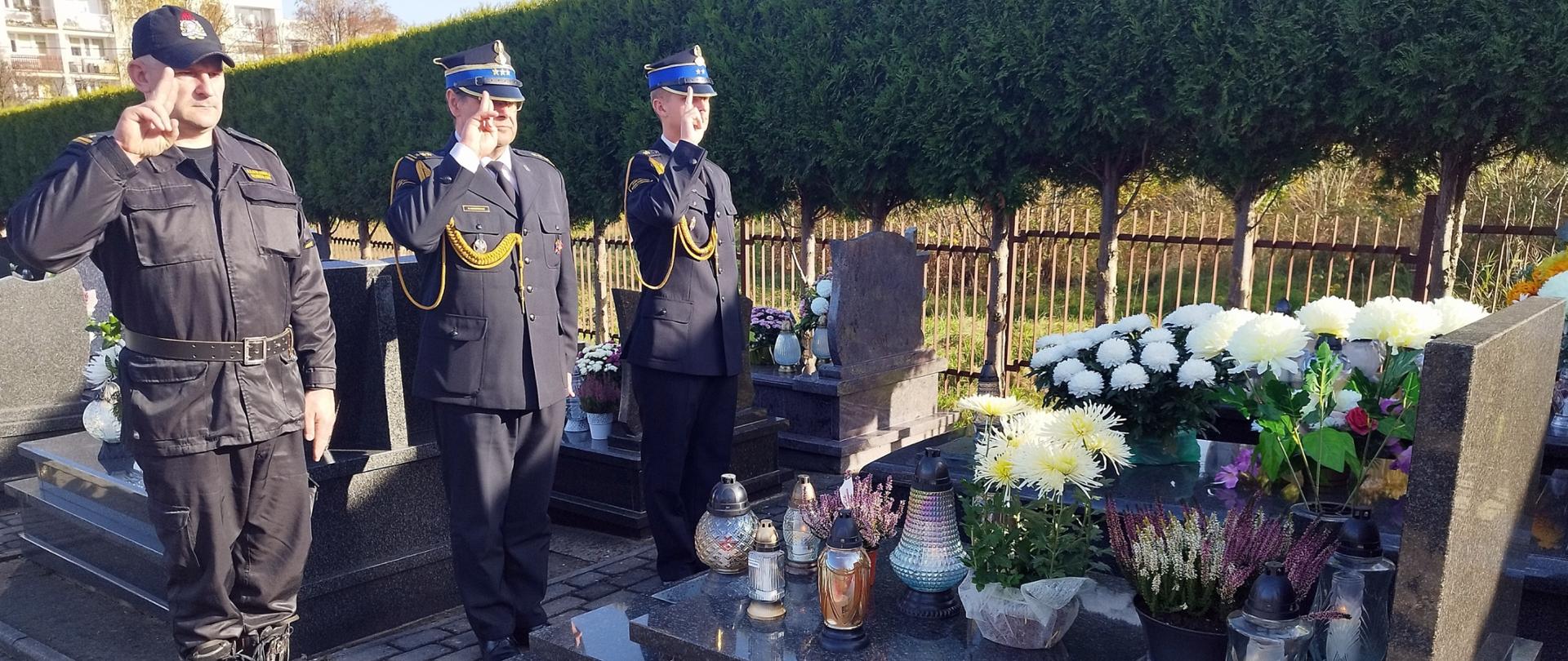 Widok z boku. Trzech strażaków PSP salutuje na cmentarzu przed grobem.