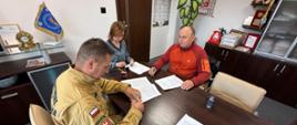 Podpisanie umowy w Komendzie Powiatowej PSP w Olecku – na zdjęciu trzech uczestników wydarzenia. Komendant PSP w mundurze, obok kobieta w sukience, mężczyzna w czerwonej bluzie. Wszyscy uczestnicy trzymają długopisy w ręce. Komendant i księgowa w trakcie parafowania stron.