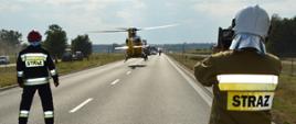 Droga krajowa nr 36. Na jezdni stoi dwóch strażaków. Obydwaj odwróceni plecami. Przed nimi (tyłem do strażaków) ląduje śmigłowiec Lotniczego Pogotowia Ratunkowego. Przed śmigłowcem widoczny rząd stojących samochodów. Z lewej strony, za siatką ogrodzeniową widoczne dwa ciągniki rolnicze oraz dwie osoby w pomarańczowych uniformach. W oddali las.