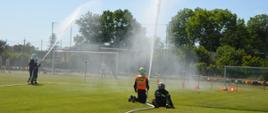 Zawody sportowo-pożarnicze. Płyta boiska na stadionie sportowym. Jedna z drużyn w końcowej fazie ćwiczenia bojowego. Podają strumienie wody w górę. Jedez zawodników znajdujących sie po prawej stronie, klęczy na ob kolanach, drugi - siedzi na murawie. W tle przewrócone pachołki oraz drezwa i krzewy