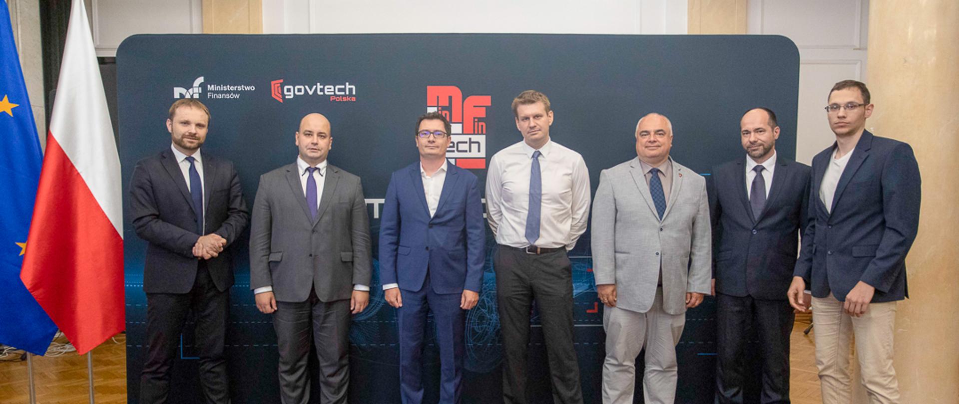Zastępca Szefa KAS Tomasz Słaboszowski wraz ze współpracownikami na tle banera MF i GOV TECH