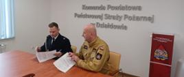 Przy stole siedzi komendant powiatowy Państwowej Straży Pożarnej w Działdowie oraz jeden ze strażaków-ochotników. Przeglądają kartki ze spisaną umową na pełnienie funkcji kierowcy w Ochotniczej Straży Pożarnej 