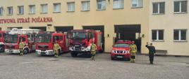 Na zdjęciu widać ustawione przed garażami samochody pożarnicze z włączonymi sygnałami świetlnymi oraz ustawionych z boku samochodów strażaków w ubraniu specjalnym.