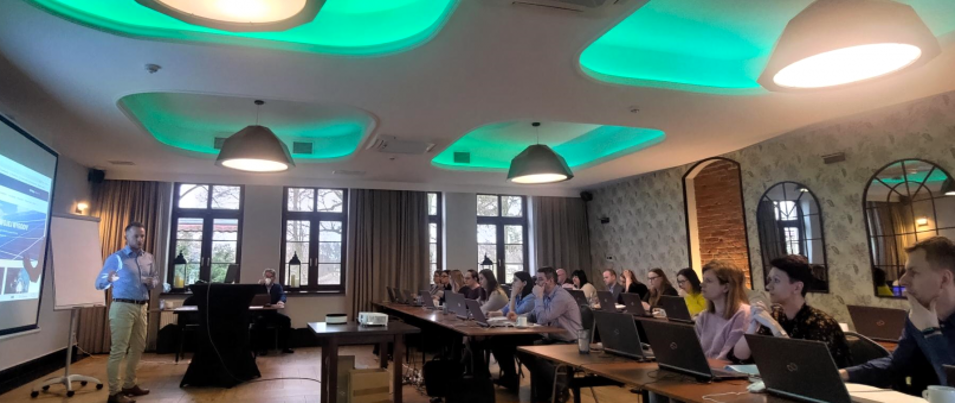 Sala konferencyjna z uczestnikami przy laptopach, prezenterem, ekranem prezentacyjnym i zielonymi akcentami oświetlenia.