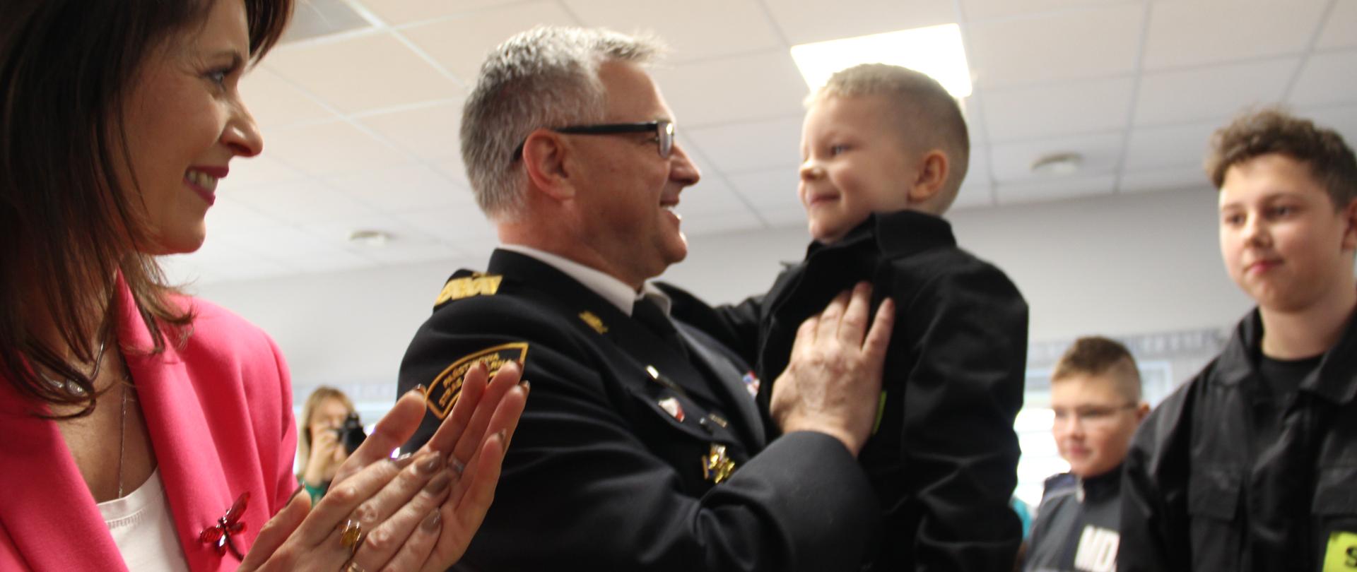 Komendant wojewodzki, w mundurze wyjściowym, trzyma na ręku kilkuletniego chłopca w mundurku strażackim. Wokół zgromadzeni goscie uroczystości.