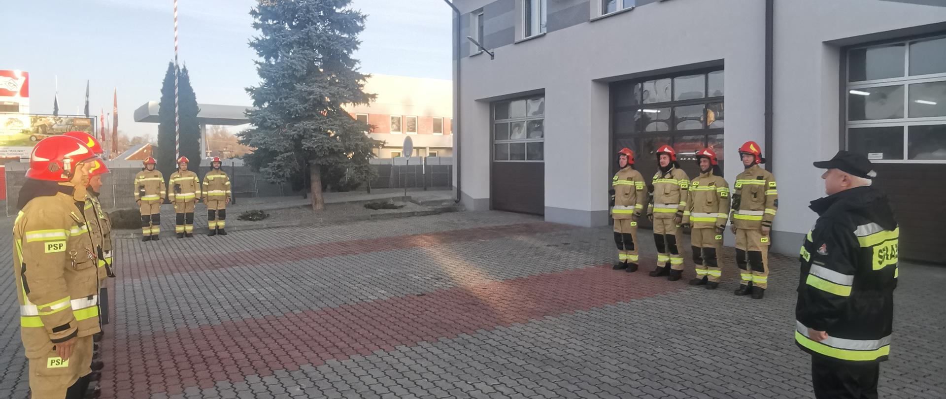 Zdjęcie przedstawia uroczystą zbiórkę z okazji Dnia Odzyskania Niepodległości. Po lewej i prawej stronie zdjęcia widoczni strażacy w ubraniach specjalnych koloru żółtego, którzy stoją w szeregu. W centralnej części zdjęcia trzech strażaków stoi przed masztem na którym wisi flaga Państwowa