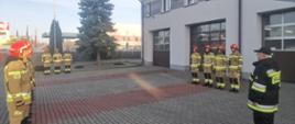 Zdjęcie przedstawia uroczystą zbiórkę z okazji Dnia Odzyskania Niepodległości. Po lewej i prawej stronie zdjęcia widoczni strażacy w ubraniach specjalnych koloru żółtego, którzy stoją w szeregu. W centralnej części zdjęcia trzech strażaków stoi przed masztem na którym wisi flaga Państwowa