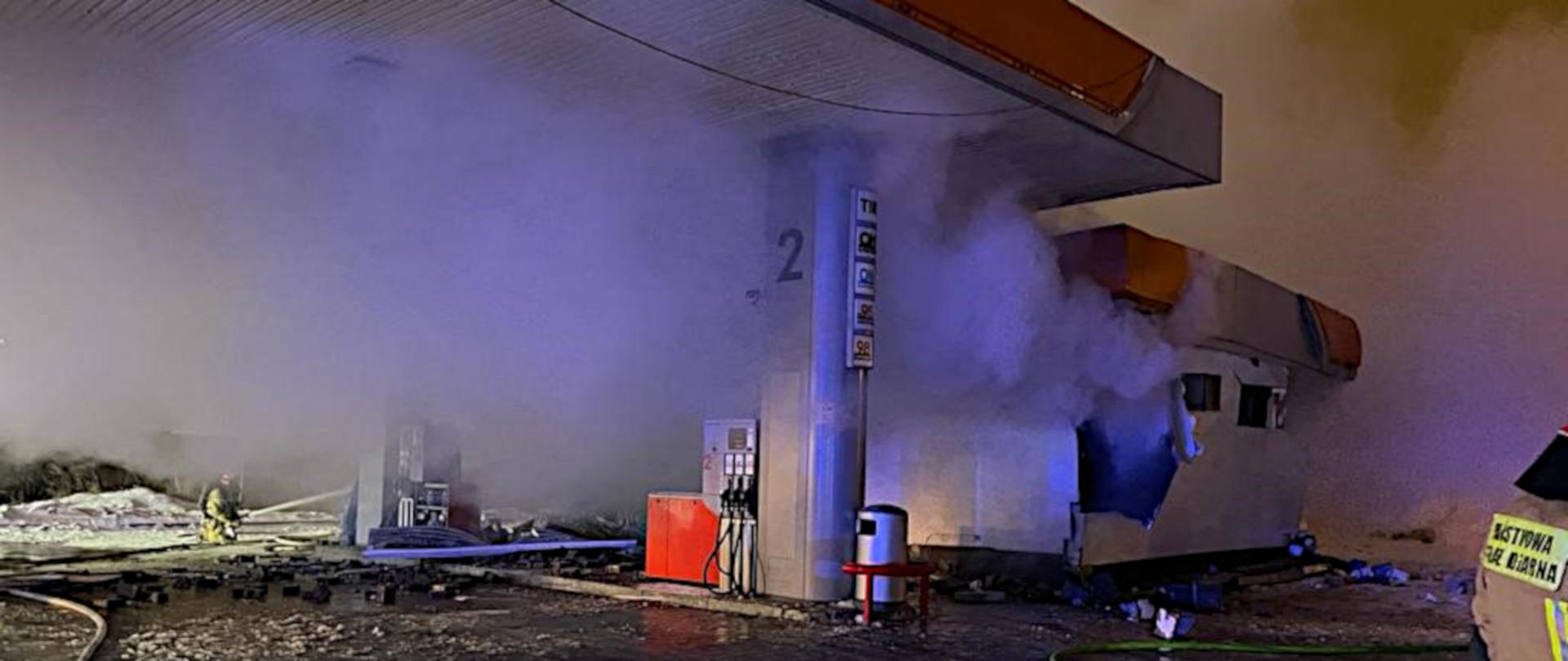Zdjęcie przedstawia stację paliw i widoczne uszkodzenia budynku na skutek wybuchu. W tle widoczny strażaka w ubrani specjalnym i hełmie, gaszący pożar z prądownicy.