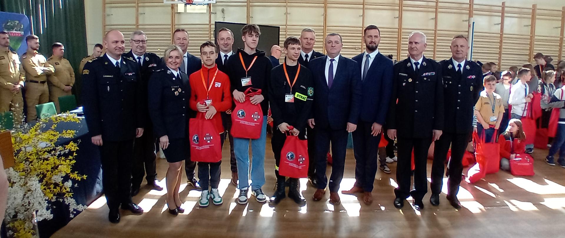 Na zdjęciu widać grupę osób dorosłych i nastolatków pozujących do zdjęcia w dużej sali gimnastycznej.
W centrum, obok siebie stoją, z lewej do prawej:
Kobieta w ciemnym mundurze z białymi akcentami i czerwoną opaską na ramieniu.
Młody mężczyzna w niebieskich dżinsach i czarnej bluzie z kapturem, trzymający czerwoną torbę.
Młody mężczyzna w jasnych dżinsach i czarnej bluzie z napisem "STRAŻ" na białej opasce, trzymający czerwoną torbę.
Mężczyzna w granatowym garniturze z niebieskim krawatem.
Mężczyzna w granatowym garniturze z granatowym krawatem.
Po bokach tej centralnej grupy stoją mężczyźni w ciemnych, galowych mundurach strażackich.
Po lewej stronie kadru widać częściowo ucięte kompozycje z żółtych kwiatów. W tle, po prawej stronie, widać grupę dzieci. Sala gimnastyczna ma drewnianą podłogę, duże okna na górze ścian i drabinki gimnastyczne na dalszej ścianie.