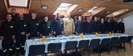 
Zdjęcie przedstawia dużą grupę strażaków stojących w szeregu w przestronnym pomieszczeniu z drewnianym sufitem, prawdopodobnie podczas spotkania wielkanocnego. W centralnej części kadru widoczne są dwa długie stoły pokryte białymi obrusami, zastawione potrawami i napojami.
Po lewej stronie stołów stoi grupa strażaków w ciemnych mundurach służbowych z żółtymi elementami. W środku kadru, za stołami, stoi mieszana grupa strażaków w mundurach oraz osób cywilnych. Wśród nich jest dwóch mężczyzn w piaskowych mundurach polowych, a także kilka osób w ubraniach cywilnych, w tym kobiety. Po prawej stronie zdjęcia, na dalszym planie, widać kolejną grupę strażaków w galowych mundurach. Na końcu pomieszczenia, na ścianie, zawieszony jest biały ekran projekcyjny.
