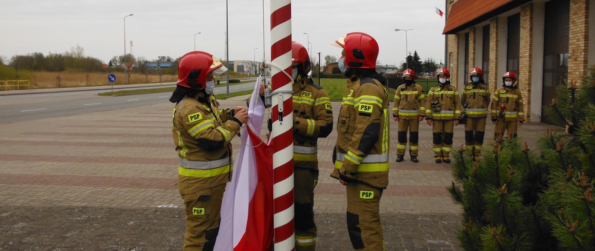 Trzech strażaków mocuje flagę państwowa do masztu, W oddali strażacy ustawieni w rzędzie wzdłuż bram garażowych