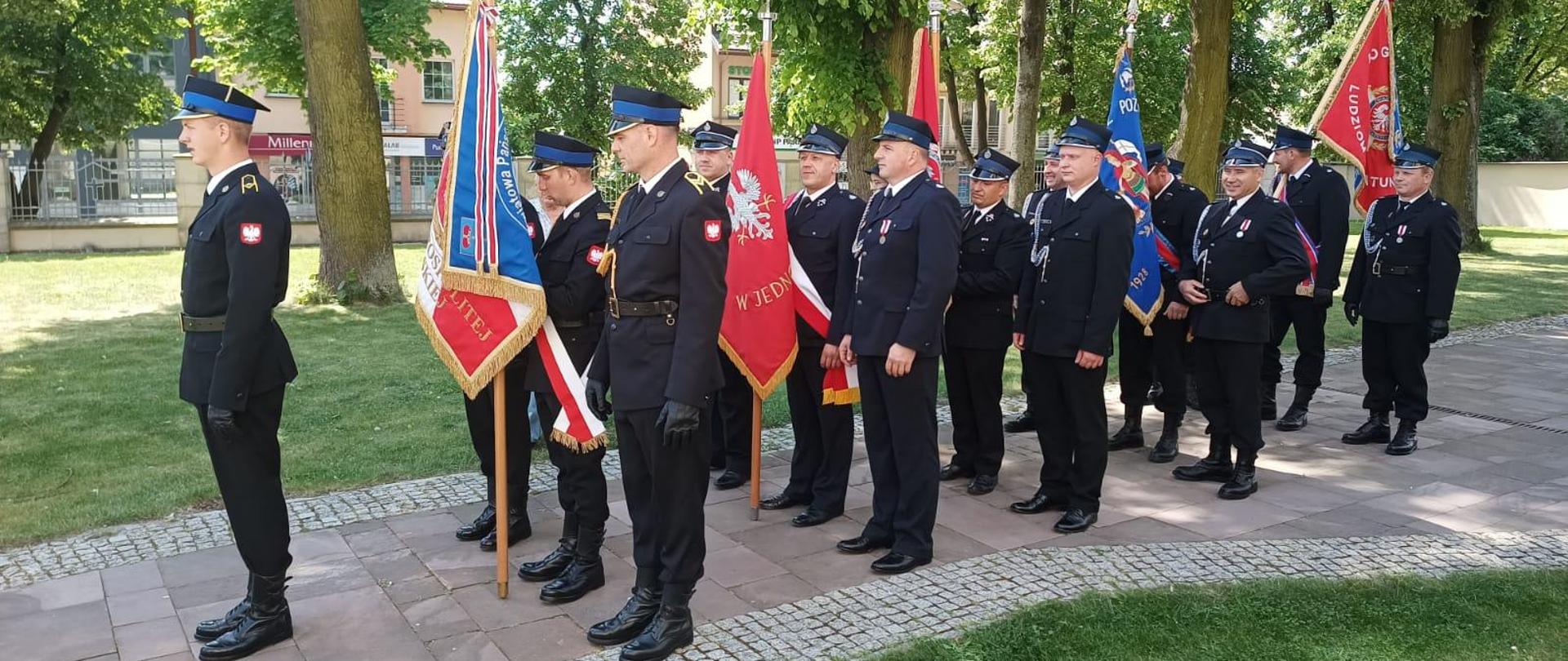 Strażacy stojący bokiem w mundurach galowych ze sztandarami, Z przodu dowódca uroczystości. W tle drzewa i budynki 