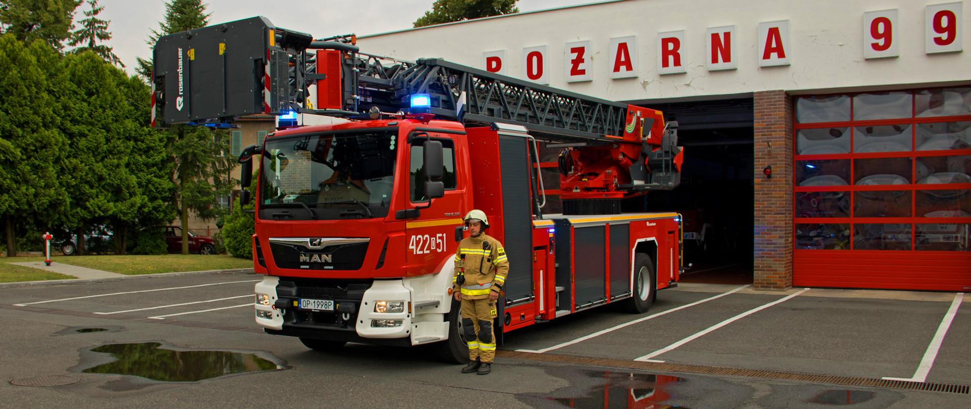 Samochód strażacki drabina a obok niej stoi strażak w tle budynek jednostki