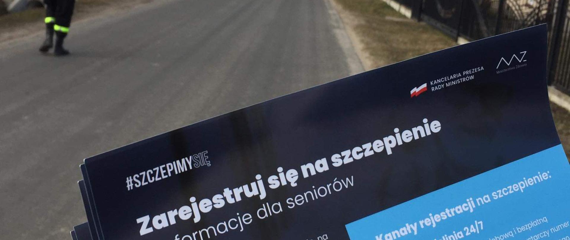 Zdjęcie przedstawia widok miejscowości. Po lewej i po prawej stronie widać prywatne posesje. Po środku znajduje się droga. Drogą wzdłuż posesji poruszają się strażacy roznoszący ulotki informacyjne o szczepieniach. 