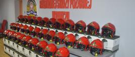 Nowe hełmy strażaków. Świetlica KP PSP w Rawiczu. Na stołach ułożone są nowe hełmy, zakupione dla strażaków JRG. Hełmy maja kolor czerwony. W tle - na ścianie - logo PSP i napis Komenda Powiatowa Państwowej Straży Pożarnej w Rawiczu. 