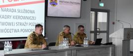 Trzech strażaków w mundurach barwy musztardowej siedzi przy biurku na którym ustawione są butelki z wodą mineralną szklanki oraz mikrofony za nimi jest ekran na którym jest wyświetlane narada służbowa kadry kierowniczej Państwowej Straży Pożarnej województwa pomorskiego obok biurka stoi mównica na meblach z przodu jest logo Państwowej Straży Pożarnej.
