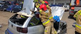 Na zdjęciu widać strażaków odginających dach samochodu osobowego do tyłu po wcześniejszym przecięciu go w odpowiednich miejscach i odcięciu od słupków pojazdu. W tle widać budynek autozłomu oraz samochodu przeznaczone do złomowania.