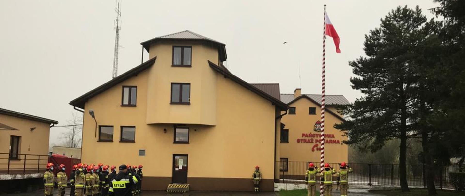 Na zdjęciu widzimy plac apelowy w KP PSP w Myślenicach. Na pierwszym planie stoją od lewej pododdział strażaków JRG, Dowódca prowadzący uroczystą zmianę służby, strażacy pocztu flagowego. W tle budynki Komendy Powiatowej PSP.