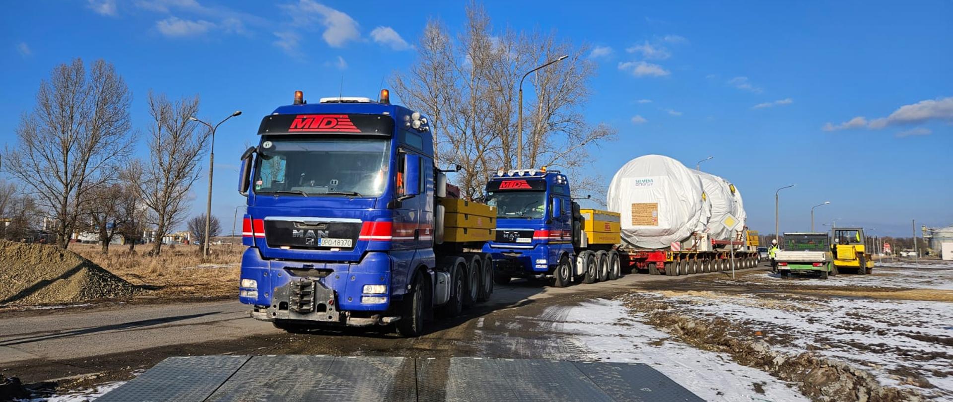 Na zdjęciu widać dwa niebieskie ciężarówki z czerwonymi i żółtymi elementami, oznaczone logo "MD". Ciężarówki przewożą duże, owinięte białą folią ładunki na wieloosiowych naczepach. W tle widać przemysłowe zbiorniki oraz kilka osób w żółtych kamizelkach odblaskowych, które wydają się nadzorować operację transportu. Na ziemi leży trochę śniegu, a niebo jest jasne i niebieskie z kilkoma chmurami.