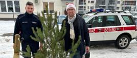 Pracownik MOPS w czapce Mikołaja i Policjant stoją z choinką i torbą z ozdobami na tle strażackiego białego Land Rovera oraz budynku mieszkalnego wielorodzinnego. Mężczyźni trzymają jeszcze w rękąch stojak do choinki i walizkę narzędziową. Na ziemi leży śnieg.