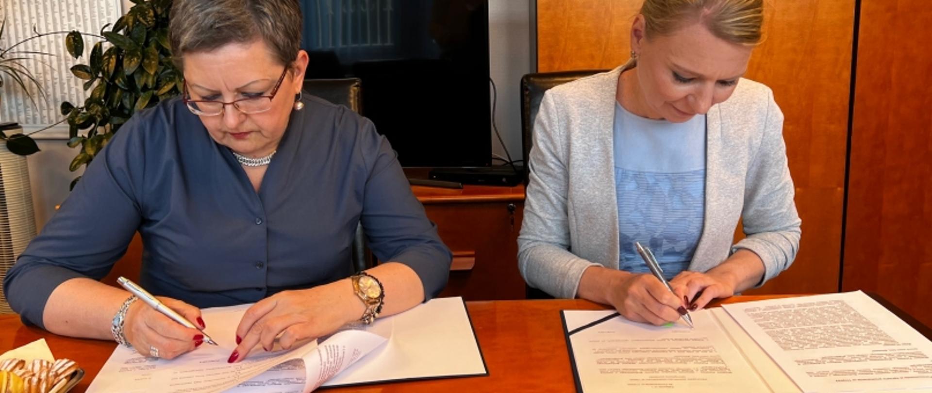 Dwie osoby podpisujące dokumenty przy stole; w tle biurowe wyposażenie i rośliny.