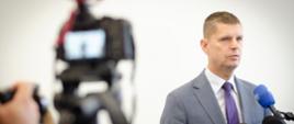 Minister stoi na tle białej ściany, przed nim mikrofony na stojakach i kamera.