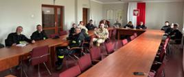 Strażacy ochotnicy uczestniczą w szkoleniu podstawowym OSOP w sali konferencyjnej. Kilkanaście osób siedzi przy ustawionych w podkowę stołach, słuchając prowadzącego i patrząc w stronę aparatu. Uczestnicy mają na sobie mundury oraz ubrania strażackie. W jasnym pomieszczeniu widać projektor sufitowy, krzesła konferencyjne oraz biało-czerwoną flagę i godło Polski na ścianie