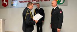 Zdjęcie przedstawia strażaków podczas wręczenia decyzji mianowania na stanowisko naczelnika wydziału operacyjno - szkoleniowego. Strażacy stoją w świetlicy