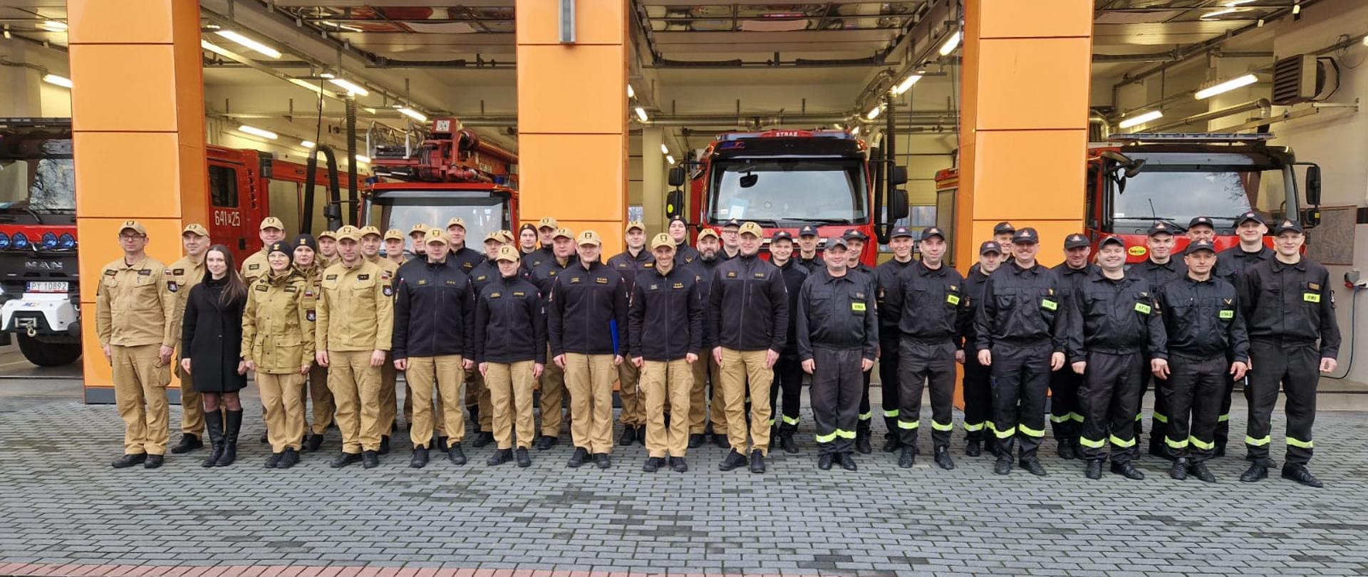 Na zdjęciu widzimy grupę strażaków ustawionych do pamiątkowej fotografii przed budynkiem remizy strażackiej. Strażacy ubrani są w mundury – część z nich ma na sobie beżowe uniformy, inni czarne ubrania ochronne z odblaskowymi elementami.
Za grupą otwarte są bramy garażowe, przez które widoczne są wozy strażackie, w tym czerwone samochody ratowniczo-gaśnicze z numerami operacyjnymi. Budynek remizy ma nowoczesny wygląd, z pomarańczową elewacją i dużymi bramami wjazdowymi.
Zdjęcie zostało wykonane po zakończeniu odprawy.
