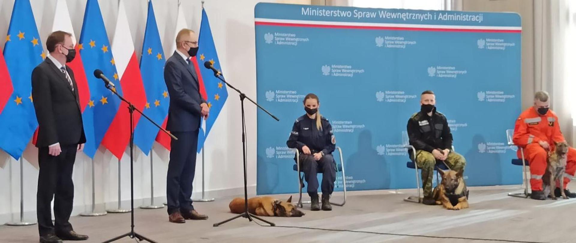 Konferencja prasowa ministra MSWiA. Na zdjęciu przedstawiciele służb ratowniczych siedzą na krzesłach a koło nich leżą psy.
