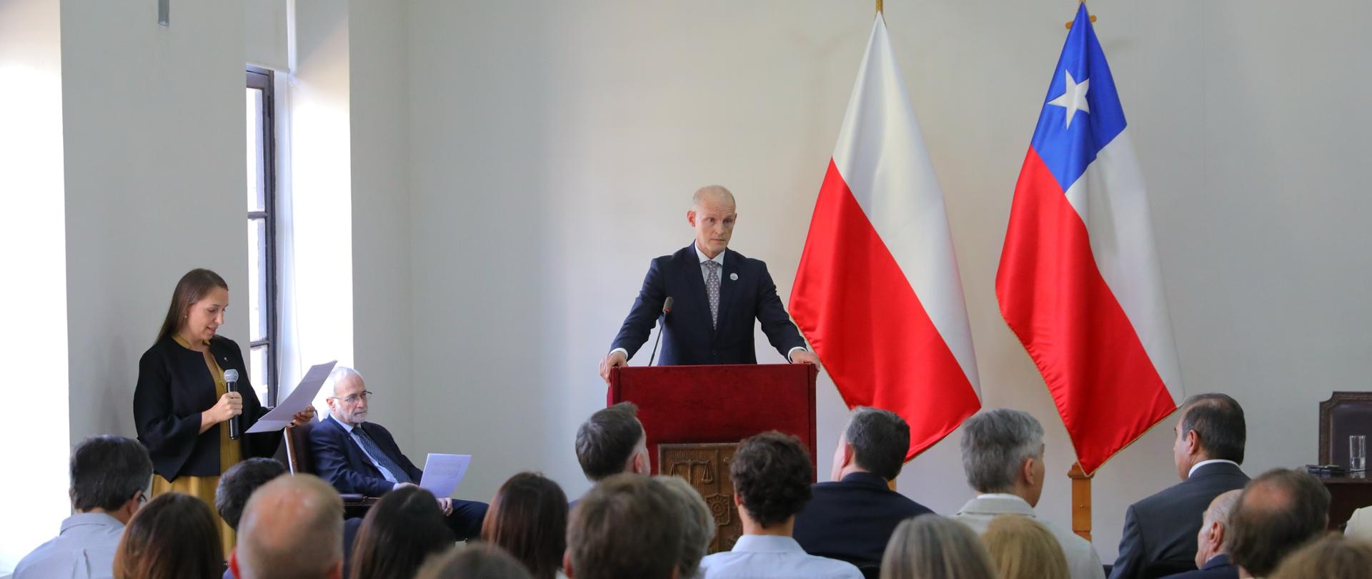 The photo shows a man in a suit standing at a lectern. Behind him are two flags: the Polish flag and the Chilean flag. To the left is a woman holding a microphone. In front of them is a group of people sitting in chairs. They have their backs to the camera. 